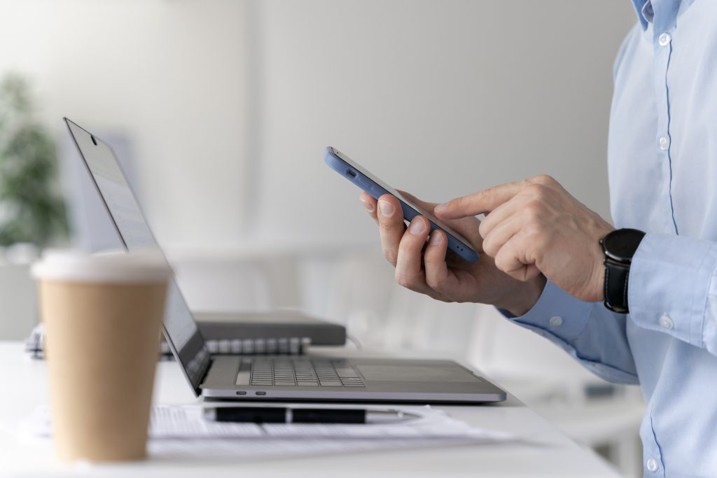 A man in the office holding his phone with a pc in front of him and a coffee on the table.